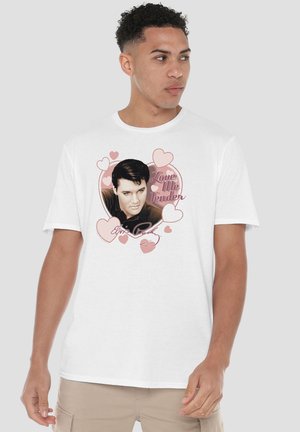 Junger Mann trägt ein weißes T-Shirt mit einem Porträt von Elvis Presley, umgeben von rosa Herzen und den Texten "Love Me Tender" und "Elvis Presley".