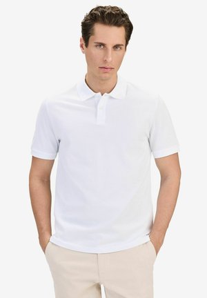 Man met lichte huid en krullend bruin haar draagt een wit poloshirt met korte mouwen en beige broek, staand met handen in de zakken.