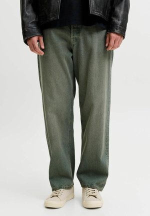 Personne portant un jean vert ample, des baskets beiges et une veste en cuir noire, debout devant un fond clair uni.
