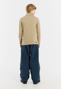 B beiger Langarmoberteil mit hohem Kragen, kombiniert mit locker sitzenden, dunkelblauen, wasserdichten Hosen. Schwarzes Akzentetikett am Saum des Shirts.