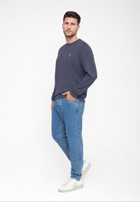 Dunkelblaue Sweatshirt mit langen Ärmeln, Rundhalsausschnitt und kleinem Logo. Kombiniert mit hellblauen Jeans und weißen Turnschuhen mit hellgrauen Akzenten.