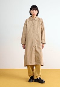 Casaco longo castanho-claro com colarinho, feito de um tecido suave. Corte solto, com bolsos laterais. Usado com calças amarelas e sapatos pretos robustos.