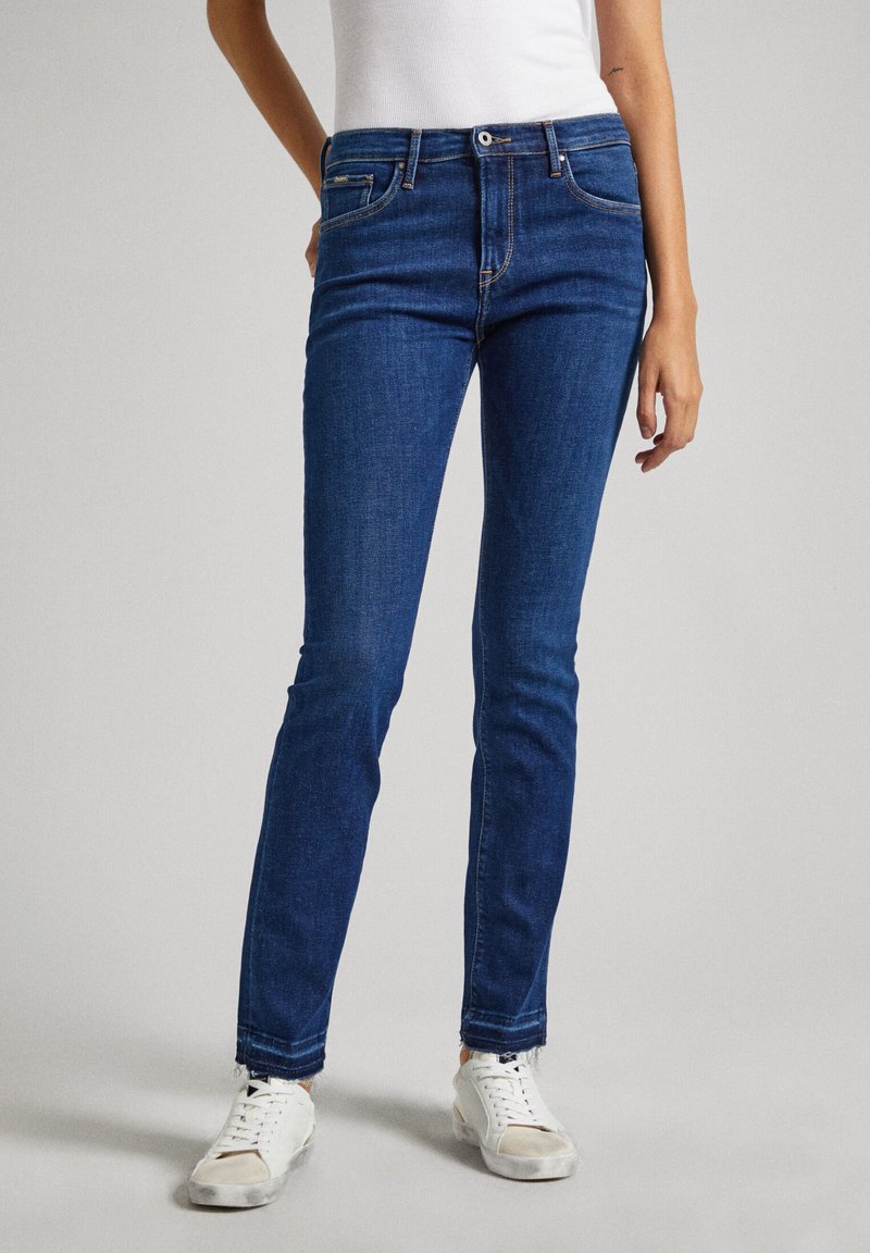 Farmernadrág, amely denim szövetből készült, slim fit szabású, öt zsebbel rendelkezik, és nyers szegéssel a bokáknál. Fehér sportcipővel párosítva.