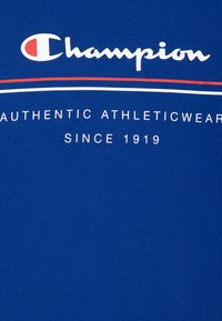 Plava pamučna majica s bijelim i crvenim natpisom te "Champion" logom s natpisom "AUTENTIČNA SPORTSKA ODJEĆA OD 1919." ispod.