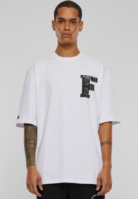 Balts, pārāk liels T-krekls no kokvilnas. Izceļas ar melnu grafiku "F" logotipu kreisajā krūtīs. Klasisks apaļais kakls un īsas piedurknes.