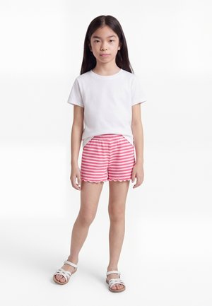 Mädchen steht und trägt ein weißes Kurzarmshirt, rosa-weiß gestreifte Shorts und weiße Sandalen vor einem schlichten weißen Hintergrund.