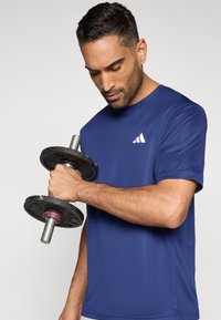 Αθλητική μπλούζα ναυτικού μπλε με κοντά μανίκια, ομαλή υφή και λευκό λογότυπο. Κρατώντας ένα μαύρο βάρος με ασημένια λαβή, δείχνοντας τον μυώδη πήχη.