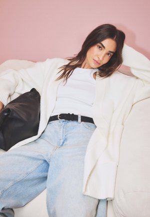 White knitted cardigan, black belt, and light blue baggy jeans. Black leather handbag with soft texture. Light background.