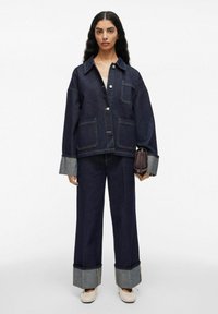 Vrouw in een oversized donkerdenim jasje en jeans met brede opgerolde manchetten, die een kleine bruine handtas vasthoudt, staande tegen een witte achtergrond.