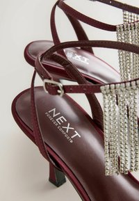 Bordeauxfarbene Riemchen-Absatzsandalen mit einem quadratischen Absatz, ausgestattet mit metallischen Fransenakzenten und einer gepolsterten Innensohle, die mit "NEXT FOREVER COMFORT" geprägt ist.