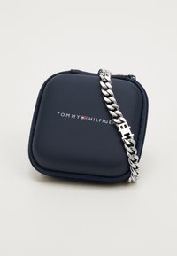 Tommy Hilfiger mornarski spremnik s patentnim zatvaračem, s srebrnim narukvicom od lančića koja ima ugravirani logo na kopči na vrhu. Glatka tekstura.