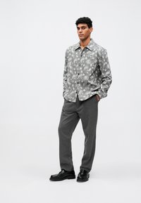 Mees seisab käed taskus, seljas mustriline pikkade varrukatega särk, hallad püksid ja mustad mokassiinid lihtsal valgel taustal.