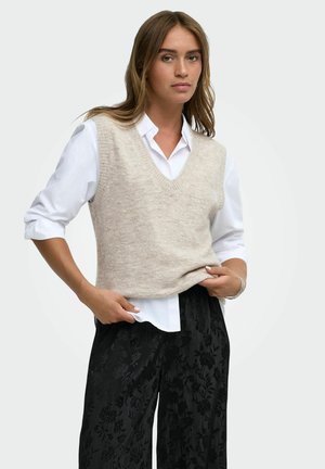 Femme portant un gilet en tricot sans manches beige sur une chemise blanche aux manches retroussées et un pantalon noir à motifs floraux.