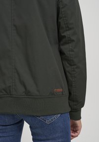 Dark green jacket with a smooth finish, ribbed cuffs and hem, and a small brown logo tag on the back. Worn with blue jeans.