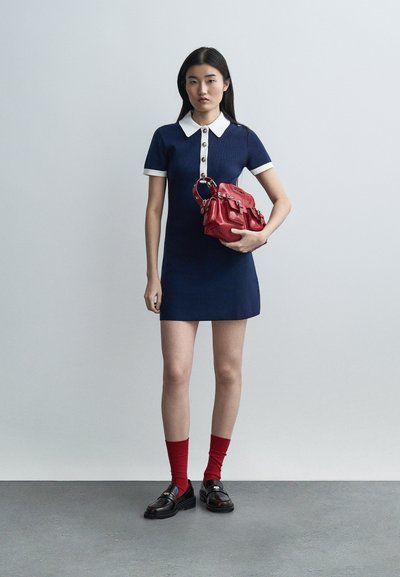 Young woman in navy dress with white collar and cuffs, holding red handbag, wearing red socks and black loafers, standing against gray background.