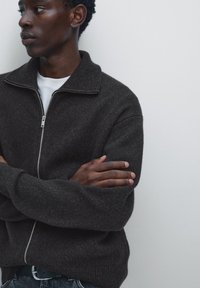 Dark gray zip-up sweater with ribbed collar. Sweater has a soft texture and relaxed fit. Model has arms crossed, wearing a white shirt underneath.
