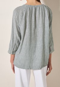 Femme portant un chemisier ample à rayures verticales vertes et blanches avec des manches aux trois quarts et un pantalon blanc, vue de dos.