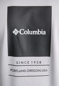 Logo Columbia sur carré noir avec texte « Depuis 1938 » et « Portland, Oregon, USA » sur fond de tissu gris clair.