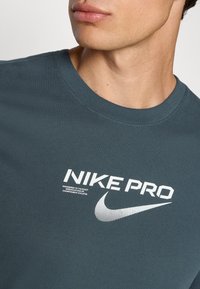 Tamsiai pilkas Nike Pro marškinėliai su trumpomis rankovėmis, apvaliu kaklu, su balta tekstu ir taškine swoosh logotipu priekyje. Minkštas audinio tekstūra.