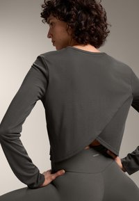 Nainen, jolla on kihara hius, pukeutuneena tummanharmaaseen pitkään hihaan olevaan toppiin, jossa on takana ylikäyvä yksityiskohta, ja yhteensopiviin korkea vyötäröön meneviin leggingseihin.