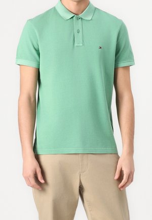Homme portant un polo vert clair à manches courtes avec deux boutons et un pantalon beige, debout devant un fond blanc uni.