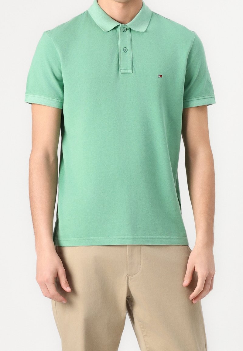 Homme portant un polo vert clair à manches courtes avec deux boutons et un pantalon beige, debout devant un fond blanc uni.