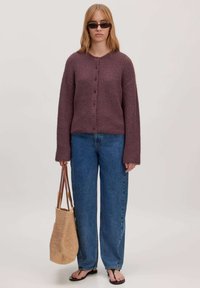Paarse gebreide cardigan met een ronde halslijn en knoppen, gecombineerd met losse blauwe jeans. Een geweven tote bag en zwarte teenslippers maken de look compleet.