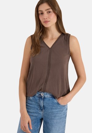 Frau mit langen braunen Haaren, die ein braunes ärmelloses Top mit V-Ausschnitt und blaue Jeans trägt, mit einer Hand in der Tasche vor weißem Hintergrund.