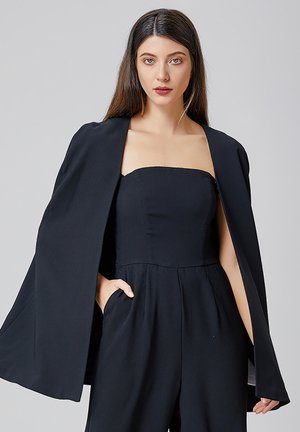 Combinaison noire avec un design sans bretelles, présentant un buste ajusté et des pantalons à jambes larges. Une cape noire assortie recouvre les épaules.