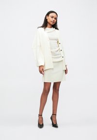 Γυναίκα που στέκεται φορώντας ένα κρεμ ζακέτα με μαύρα κουμπιά, μια κρεμ φούστα και μαύρα ψηλοτάκουνα παπούτσια μπροστά σε λευκό φόντο.