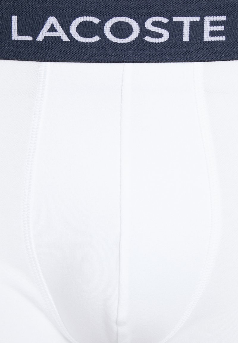 Weiße Baumwoll-Boxershorts mit einem marineblauen elastischen Bund, der den Markennamen "Lacoste" in weißer Schrift trägt. Glatte Textur und enganliegendes Design.
