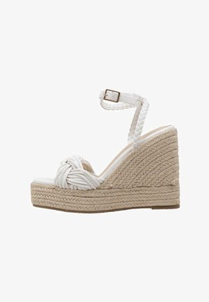 Sandalia de cuña blanca con diseño de cuerda trenzada, que presenta una correa ancha con detalle de nudo y un tacón de yute texturizado. Correa ajustable en el tobillo.