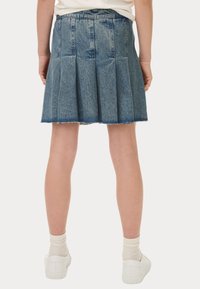 Jupe plissée en denim avec un ourlet brut, de couleur bleue, bande au niveau de la taille et coutures visibles. Portée avec des baskets blanches et des chaussettes pastels.