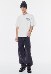 Weißes Baumwoll-T-Shirt mit grafischem Aufdruck "FIRST FLIGHT" in Blau, kombiniert mit locker sitzenden, dunkelgrauen Jeans und schwarzen Schuhen. Strukturierte Strickmütze.