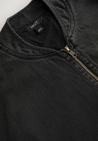 Černá manchestrová bunda se zlatým zipem částečně zapnutým, ukazující černou vnitřní podšívku a štítek značky "NEXT" na límci.