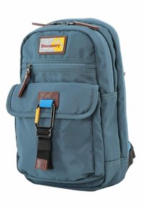 Zaino in tessuto blu con tasca frontale, dettagli marroni e una toppa rettangolare con la scritta "Discovery". Presenta zip e spallacci regolabili.