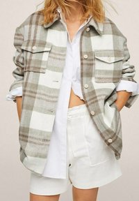 Camisa chaqueta de cuadros gris y blanco, de corte oversize, con dos bolsillos frontales, cierre de botones y cuello, superpuesta a una camisa blanca.