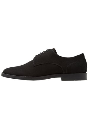Zapato de vestir de ante negro con un acabado liso, ligera curvatura en el diseño y suela de goma plana. Presenta detalles mínimos de cordones y costuras.