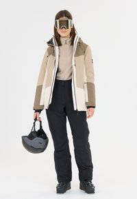 Completo da sci con giacca beige e bianca, pantaloni neri e scarponi da sci neri. Inclusi occhiali trasparenti e un casco nero.