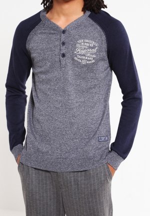 Homme portant une chemise henley grise avec des manches raglan bleu marine et un logo imprimé sur la poitrine, associée à un pantalon gris à fines rayures.