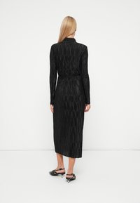 Fekete, redőzött ruha hosszú ujjal, magas nyakkal és megkötött derékkal, amely a térd közepéig ér. Fekete, nyitott orrú cipővel párosítva.