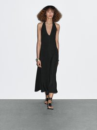 Black halter-neck dress with a deep V neckline and a flowing silhouette. Model wears black strappy sandals and a gold cuff bracelet.