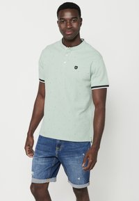 Polo shirt verde chiaro con colletto a coste, bottone a placca e piccoli accenti blu navy. Abbinato a pantaloni shorts in denim, con finitura consumata.