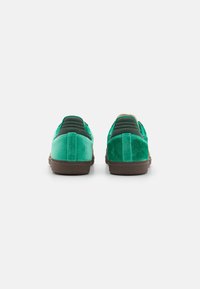 Des chaussures slip-on en velours vert avec une texture lisse, dotées d'une semelle en caoutchouc brun et d'accents rembourrés verts au niveau du talon.