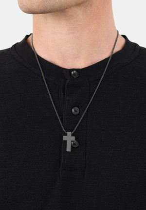 Camicia nera dal tessuto texturizzato con scollo henley e tre bottoni; un ciondolo a croce scuro su una catena d'argento pende lungo lo scollo.