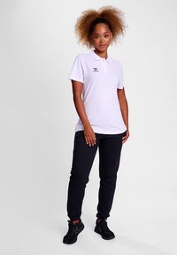 Ljuslila pikétröja med krage och broderad logotyp, kombinerad med svarta mjukisbyxor och svarta sportskor. Slät textur, avslappnad passform.