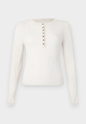 Hosszú ujjú krémszínű pulóver bordázott textúrával, kerek nyakkal és hét dekoratív gombbal az elején.