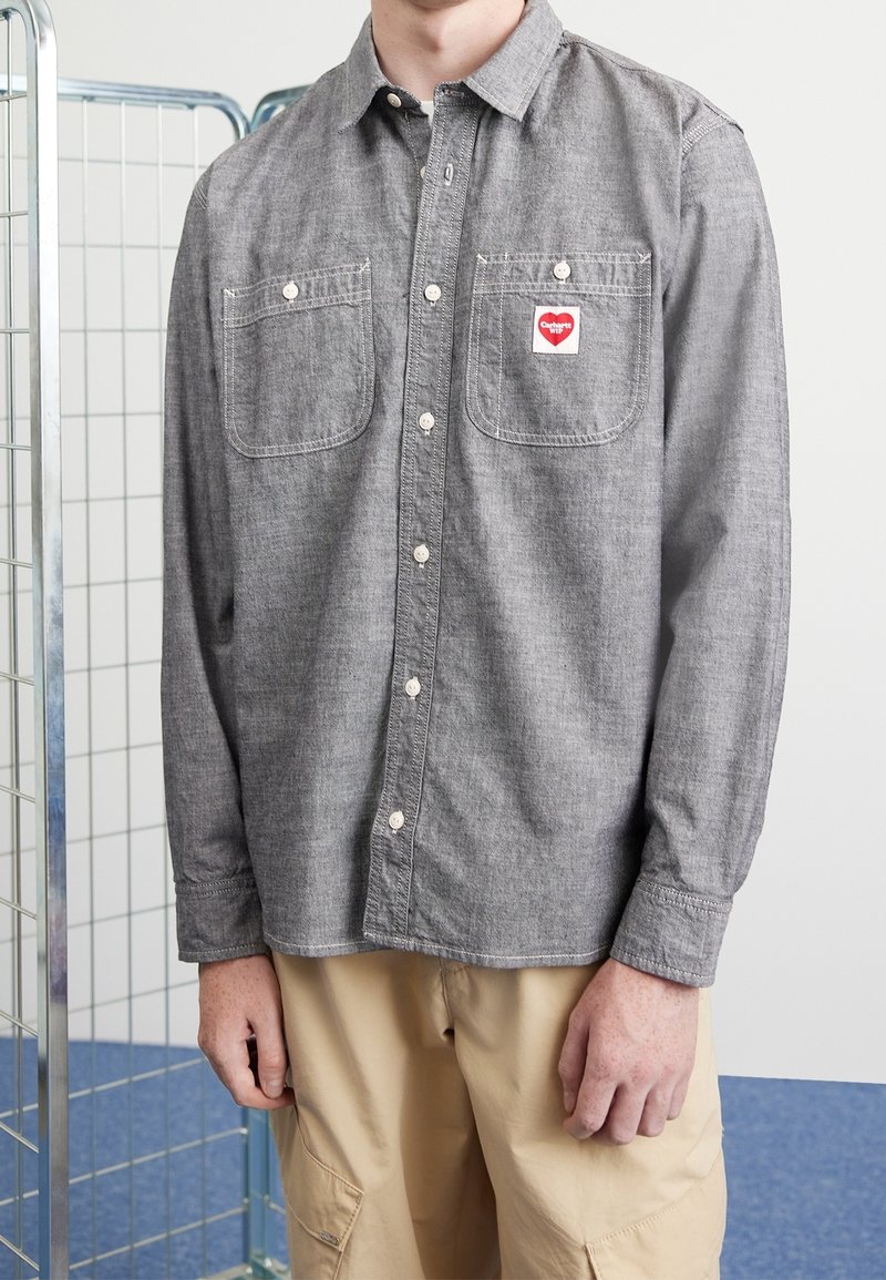 Persoon draagt een grijs overhemd met knopen en twee borstzakken, waarvan één met een rood hartlogo, en beige cargobroek.