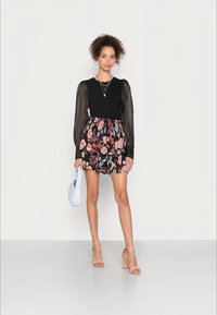 Top preto de manga longa com mangas bufantes transparentes e saia mini floral em camadas. A modelo segura uma mala azul-claro e usa sandálias de salto nu.