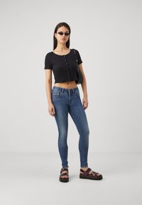 Svart kortärmad croppad topp med knapppdetaljer, blå slim-fit jeans och mörka sandaler. Modell som håller en liten svart handväska.